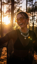 A woman wearing round dark sunglasses, layered silver necklaces, and a black mesh top stands in a sunlit forest at sunset.