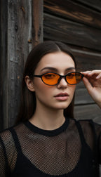 A woman wearing black-framed sunglasses with vibrant orange tinted lenses, holding one lens with her hand against a rustic dark wood background.
