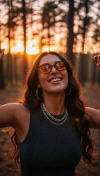 A woman outdoors in a forest at sunset wearing orange-tinted sunglasses, a dark sleeveless top, layered silver chain necklaces, and hoop earrings.