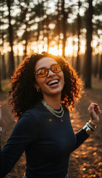 A woman with curly dark hair and orange-tinted glasses smiles joyfully in a sunlit forest at sunset, wearing a dark long-sleeve top and layered silver necklaces.