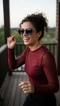 A woman with curly dark hair wearing a fitted burgundy long-sleeve top and black sunglasses, smiling on a wooden deck at dusk.