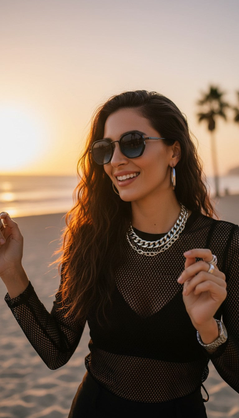 A woman wearing round sunglasses, a chunky silver chain necklace, and a black long-sleeve mesh top stands on a beach at sunset with the ocean and palm trees in the background.